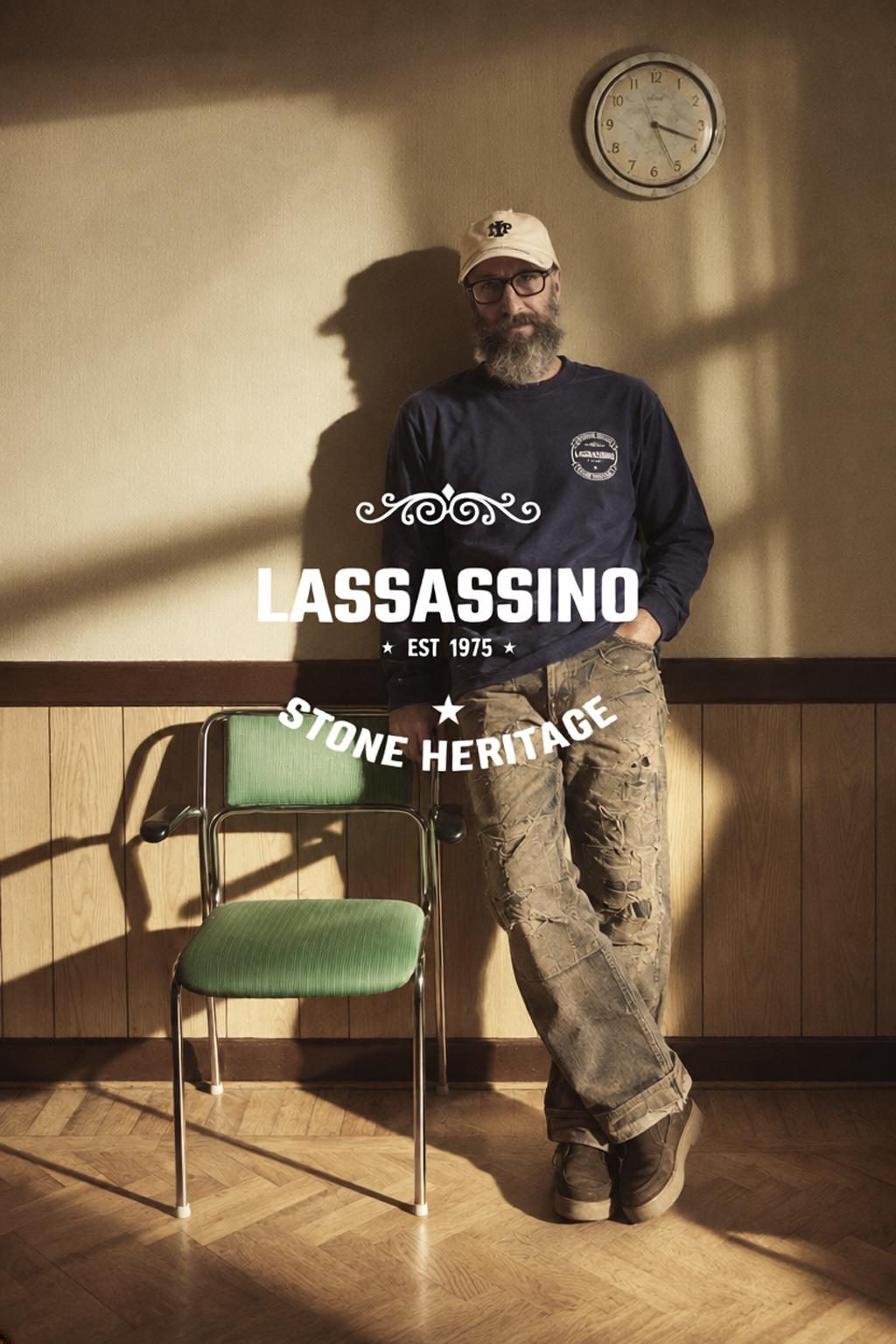 Uomo barbuto con felpa blu e jeans consumati accanto a sedia verde, scritta LASSASSINO EST 1975 Stone Heritage
